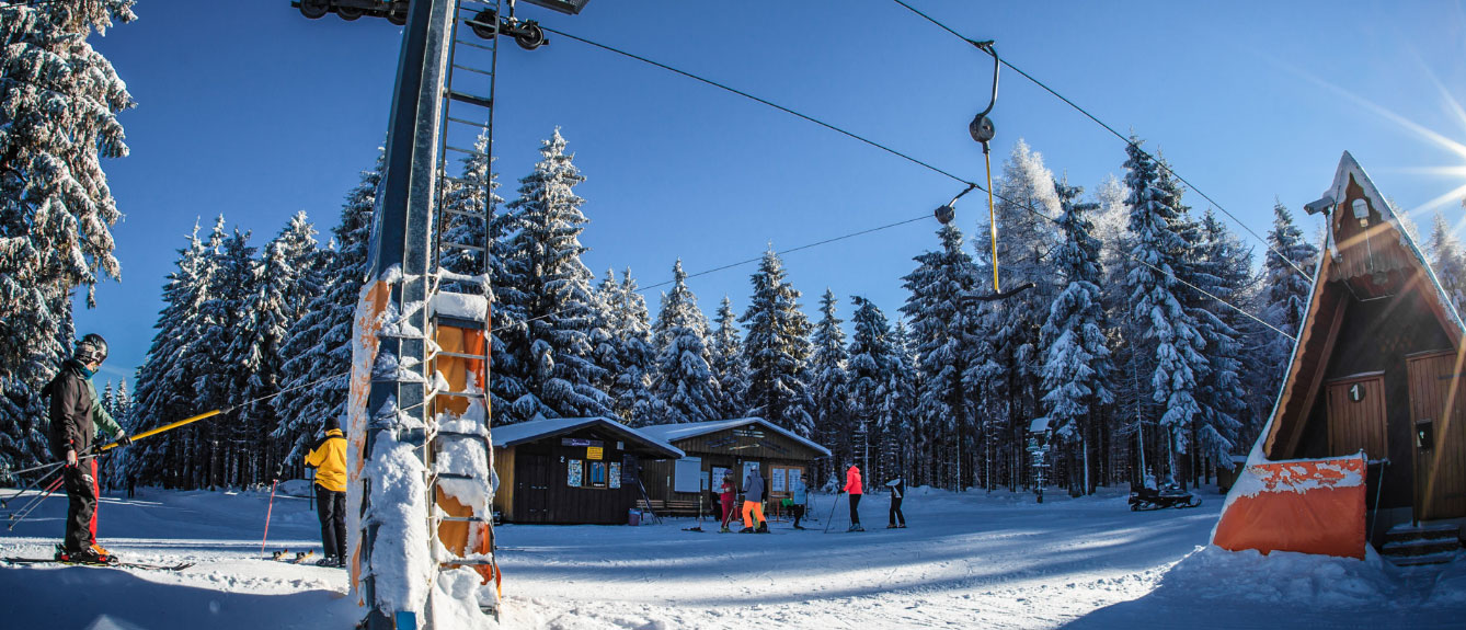 Winterlandschaft mit Skilift, Menschen auf Skiern und im Hintergrund verschneite Bäume mit Hütten
